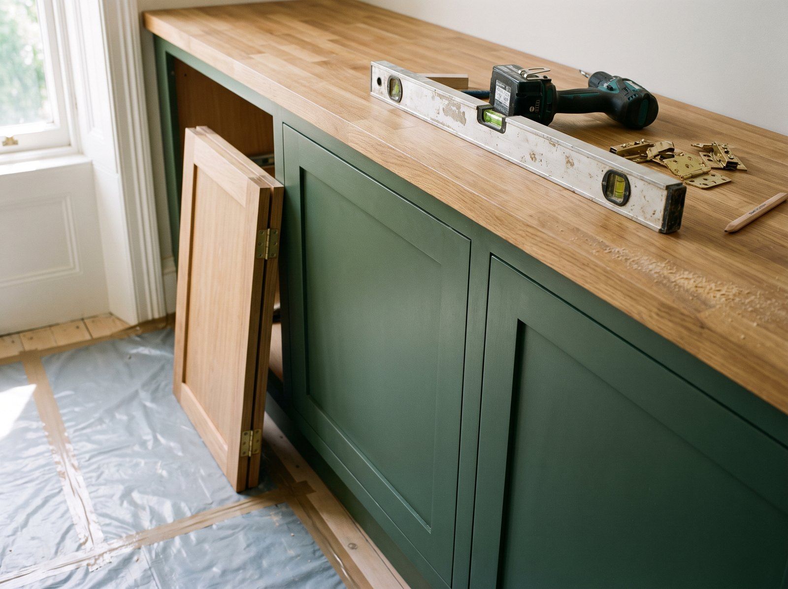 Meubles de cuisine verts en cours de pose sous un plan de travail en chêne, niveau et visseuse posés dessus — image d’illustration